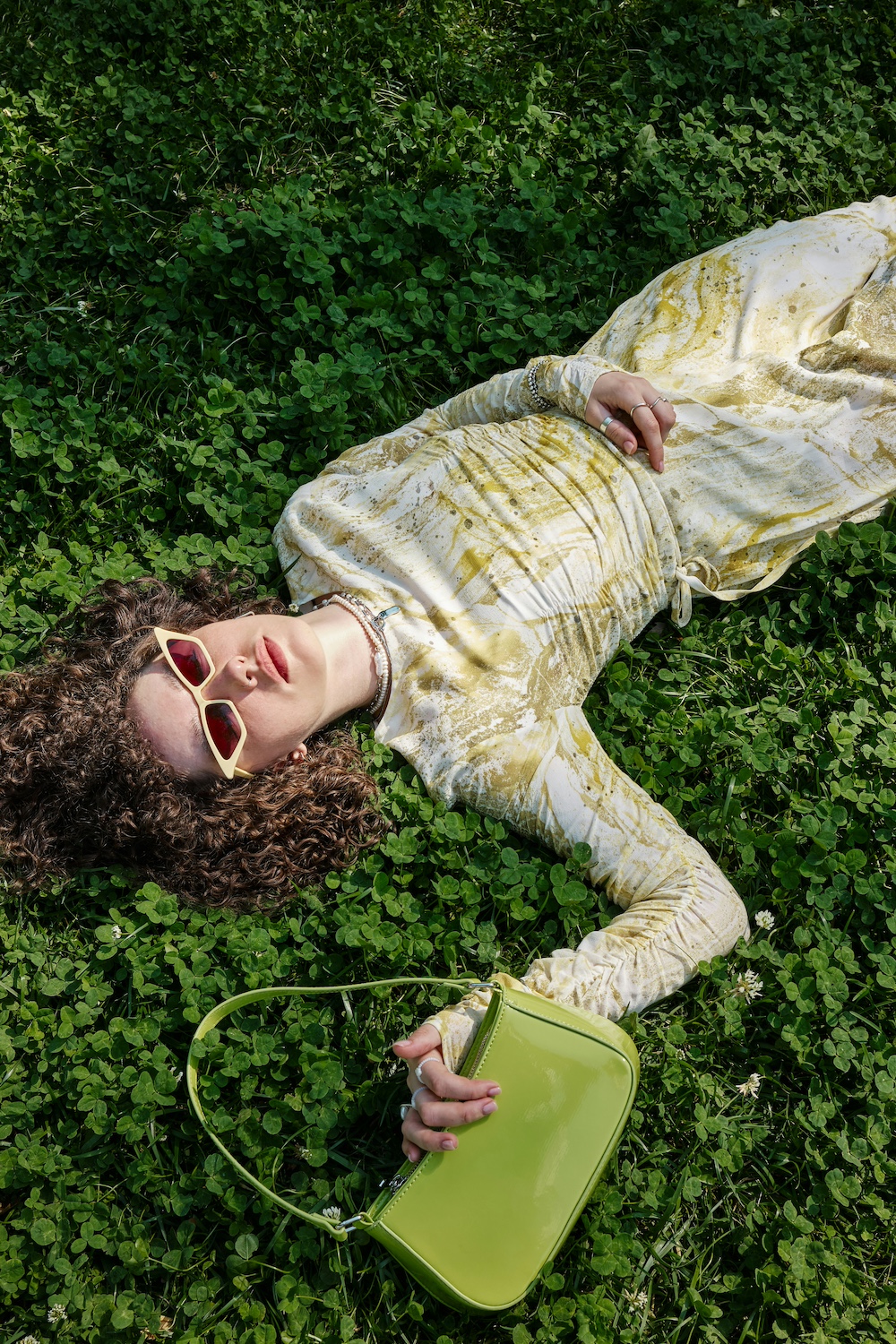 Model lying in clover grass holding a lime patent shoulder bag
